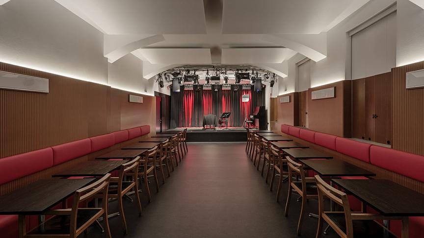 © ORF/Ö1/Hertha Hurnaus Blick in das leere ORF RadioCafe mit roten Sitzbänken und Holztischen und eine kleine Bühne mit rotem Vorhang und Beleuchtung.