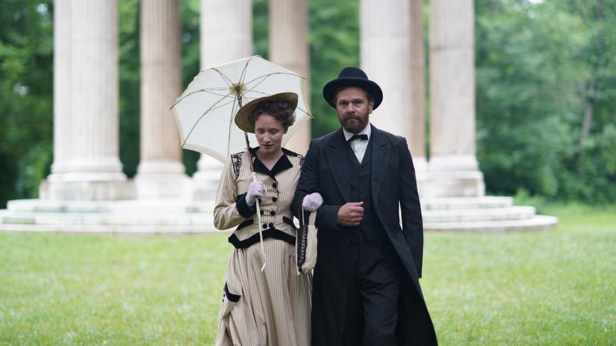 © ORF/Ranfilm Per Lasson als Alfred Nobel und Lili Winderlich als Bertha von Suttner spazieren in historischer Mode vor antiken Säulen durch einen Park.