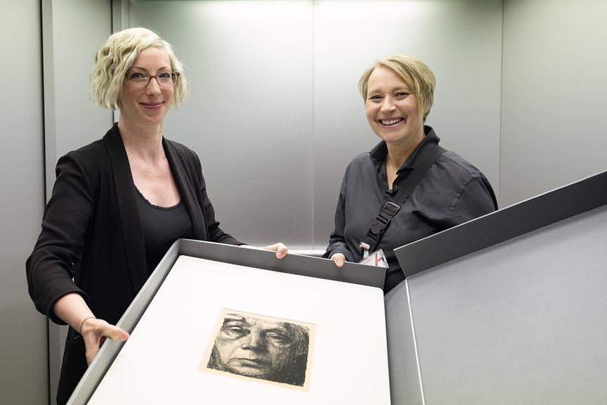 © ORF/V-set GmbH/Stefan Vucsina Regisseurin Alex Wieser und Beatrix Brunschko stehen in einem schlichten Raum und präsentieren gemeinsam eine große, geöffnete Box, in der ein gerahmtes Kunstwerk liegt.