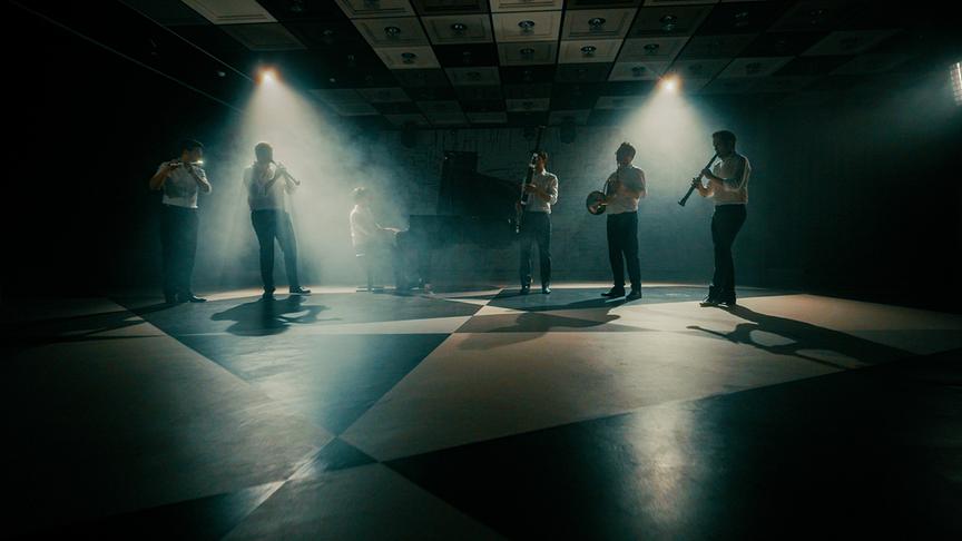 © ORF/V-set GmbH Eine Gruppe von Musikerinnen und Musikern spielt in einem stimmungsvoll ausgeleuchteten Raum verschiedene Blas- und Tasteninstrumente.