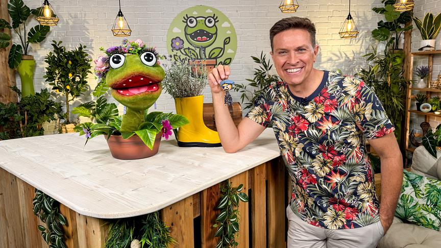 © ORF/Steiner Familyentertainment Robert steht in einem begrünten Studio an einem Holztresen neben der Pflanzen‑Puppenfigur „Lila Grasgrün“.