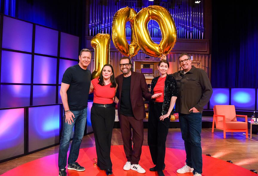 © ORF/Hubert Mican Gruppenfoto mit Alex Kristan, Ina Jovanovic, Gerald Fleischhacker, Chrissi Buchmasser und Viktor Gernot stehen im "Die Tafelrunde"-Studio auf einem roten Teppich und posieren gemeinsam vor großen goldenen Luftballons in Form der Zahl 100.