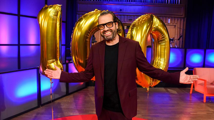 © ORF/Hubert Mican Gerald Fleischhacker steht mit ausgebreiteten Armen im "Die Tafelrunde"-Studio auf einem roten Teppich vor großen goldenen Luftballons in Form der Zahl 100.