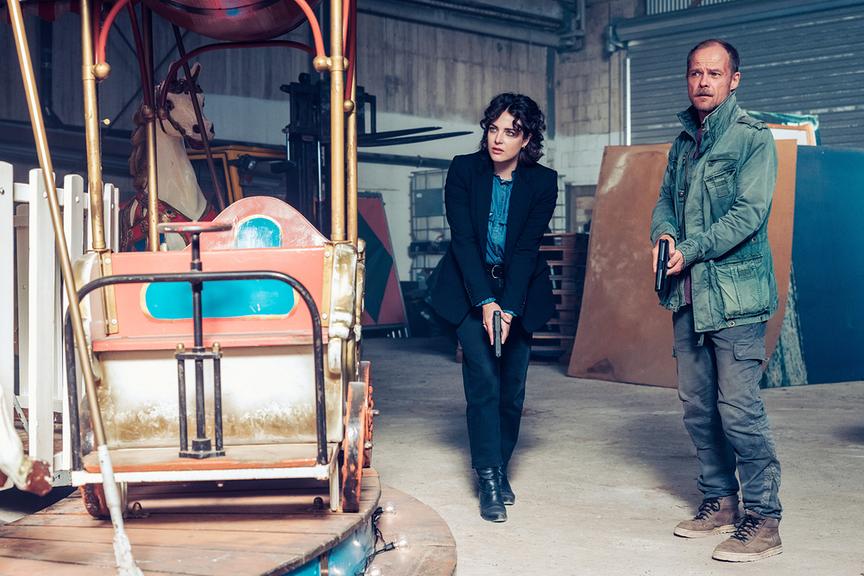 Mara Eisler und Micha Oberländer mit Pistolen in den Händen stehen in einer Lagerhalle neben einem alten Karussell.