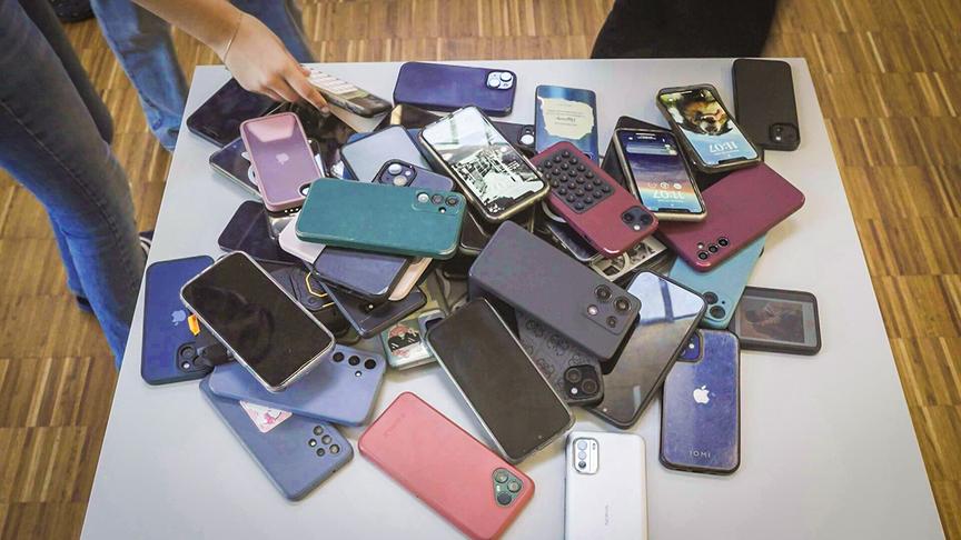 Ein großer Haufen unterschiedlichster Smartphones liegt auf einem Tisch, während Schüler:innen ihre Geräte dort ablegen oder wegnehmen.