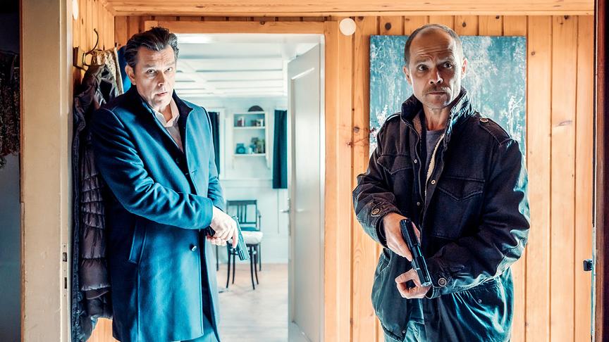 Hary Prinz und Matthias Koeberlin bei den Dreharbeiten stehen nebeneinander in einem holzgetäfelten Raum und halten jeweils eine Pistole in der Hand.