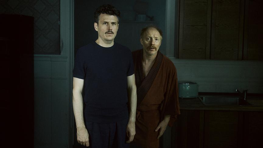 © ORF/Lotus Film/Felix Vratny Manuel Rubey als Stefan und Simon Schwarz als Theo im Morgenmantel stehen in einer düsteren, altmodisch eingerichteten Küche am Set von „Halbweg“ (AT).