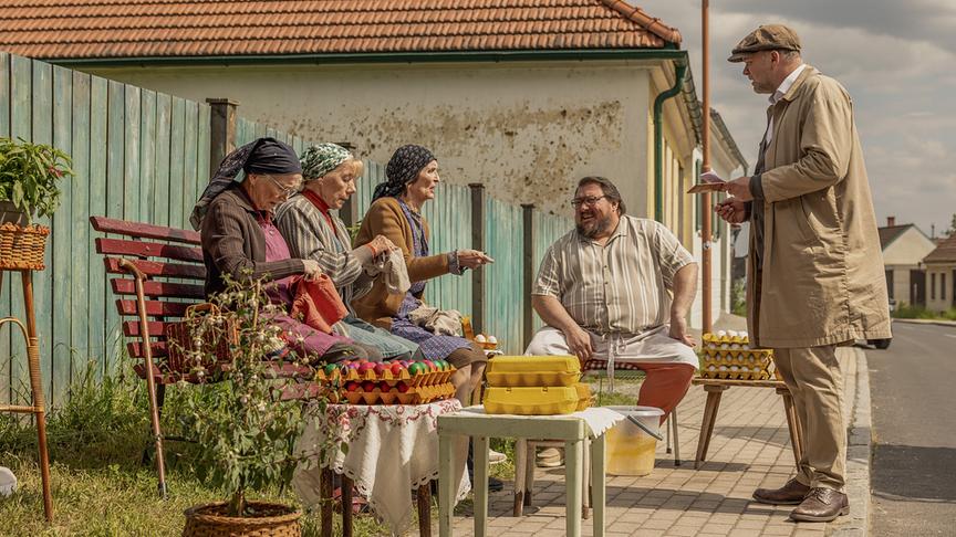 © ORF/Mona Film/Victoria Herbig Resl Grandits, Baba Sifkovits, Hilda Resetarits und Franz Maikits sitzen auf dem Gehsteig vor einem Haus an einem improvisierten Verkaufsstand mit Eierkartons, während Sifkovits vor ihnen steht und etwas notiert.