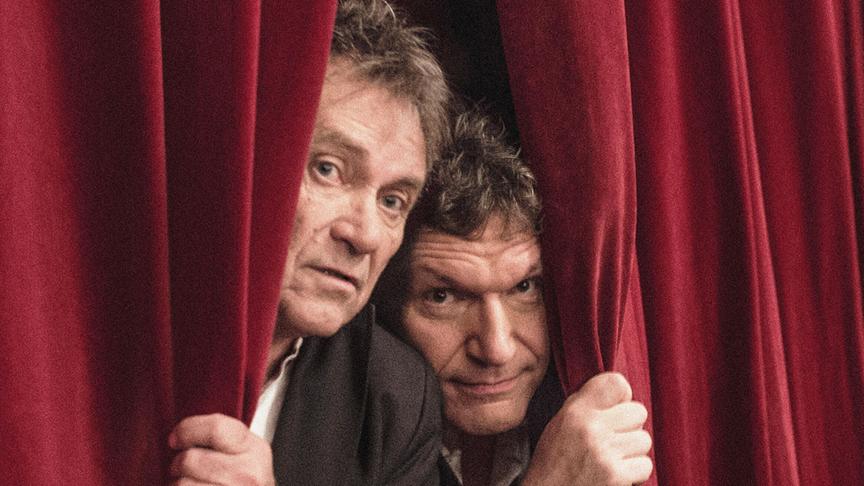 © ORF/Christian Jungwirth Wolfram Berger und Markus Schirmer schauen zwischen einem schweren roten Theatervorhang hervor.