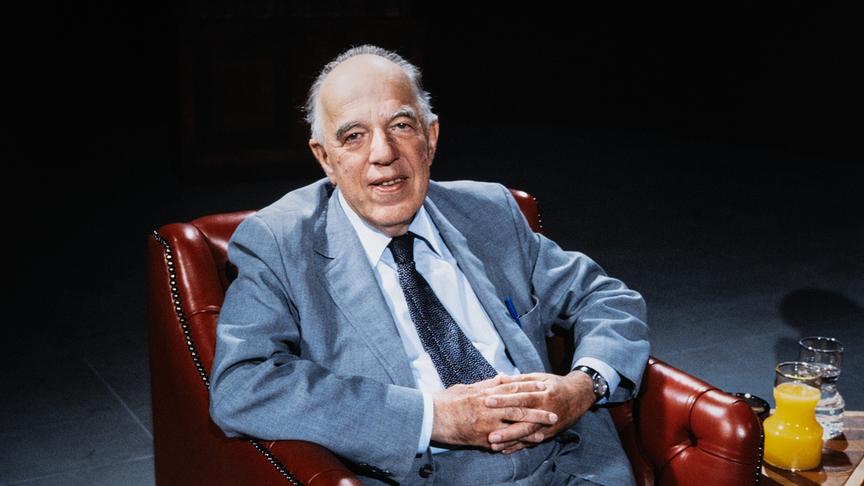 Kunsthistoriker Ernst H. Gombrich sitzt in einem roten Ledersessel vor dunklem Studioboden.