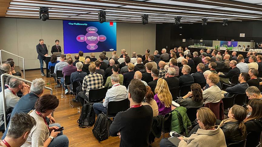 Eine große Gruppe von Menschen, die in einem modernen Konferenzraum einem Vortrag zweier Präsentierender beim ESC Production Meeting vor einer Leinwand folgen.