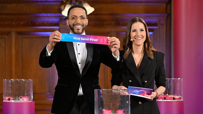 Cesár Sampson präsentiert neben Alexandra Maritza Wachter bei einer Auslosung ein farbiges Schild mit der Aufschrift "First Semi-Final" in einem festlich dekorierten Raum.