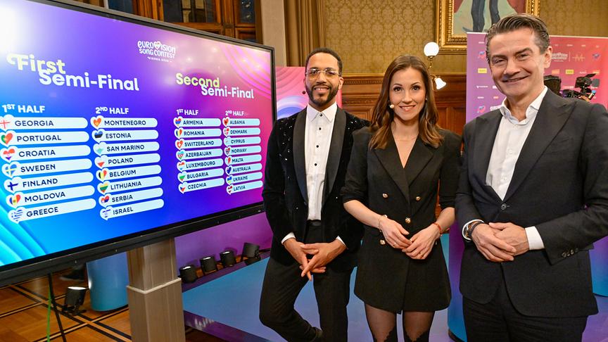 Cesár Sampson, Alexandra Maritza Wachter und ORF-Generaldirektor Roland Weißmann stehen im Wiener Rathaus neben einem Bildschirm, der die Auslosung der Halbfinals des Eurovision Song Contest zeigt.