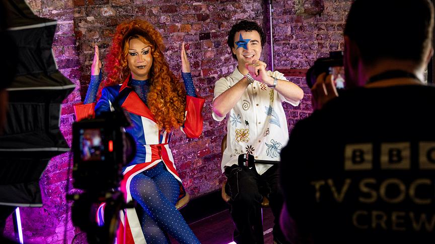 COSMÓ sitzt neben einer Frau vor einer Kamera in einem Studio‑Setting und zeigt Tanzbewegungen, während ein BBC-Kamerateam sie filmt.