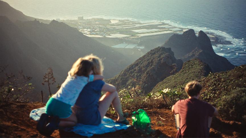 Filmszene aus "Everytime": Rückenansicht von drei Menschen, die auf einer Anhöhe sitzen und bei warmem Licht auf eine zerklüftete Küstenlandschaft mit Meer und Feldern hinabblicken.