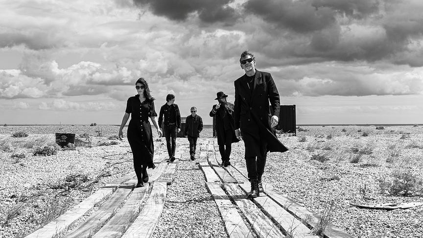 Die Band "Archive" in dunkler Kleidung geht über einen schotterigen, verlassen wirkenden Landschaftsstreifen unter stark bewölktem Himmel.