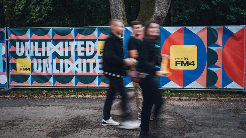 Drei verschwommen vorbeigehende Personen passieren ein buntes Banner mit der Aufschrift „Unlimited“ und dem Logo von Radio FM4.