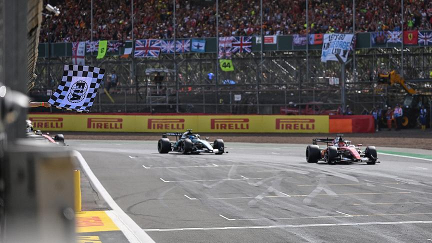© APA/AFP/POOL/PETER POWELL Zwei Formel-1-Autos mit Ferrari-Pilot Charles Leclerc im ersten Auto fahren vor voll besetzten Tribünen über die Ziellinie, während links eine schwarz-weiß karierte Flagge geschwenkt wird.