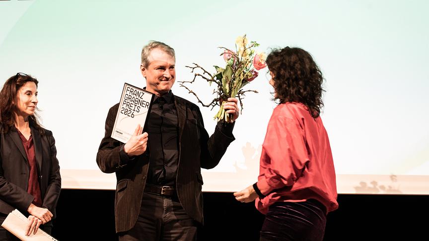 © ORF/Diagonale/Miriam Raneburger Auf einer Bühne wird bei der Preisverleihung Robert Styblo der Franz-Grabner-Preis 2026 und ein Blumenstrauß überreicht, während Karin Moser und eine weitere Frau danebenstehen.
