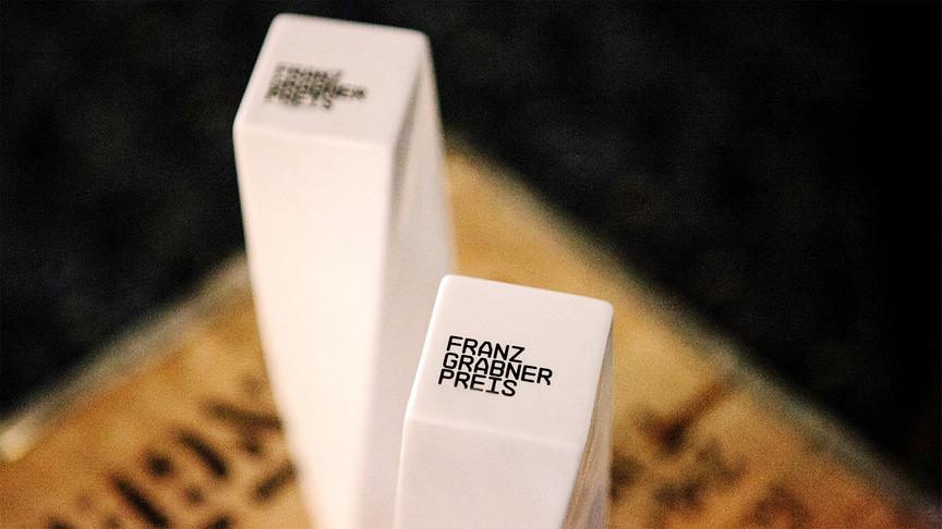 Zwei moderne, weiße, quadratische Preisstatuetten mit der Aufschrift „Franz‑Grabner‑Preis“ stehen auf einem Sockel und sind stimmungsvoll ausgeleuchtet.