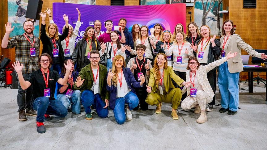 Eine große Gruppe von Teilnehmerinnen und Teilnehmern beim Volunteers-Casting posiert gut gelaunt mit erhobenen Händen vor einer farbigen Event-Bühne mit Namensbadges um den Hals.