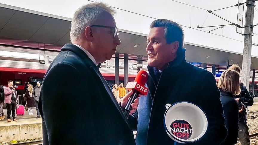 Peter Klien befragt auf einem Bahnsteig Peter Hanke, , der den weißen "Gute-Nacht-Topf" in der Hand hält, während im Hintergrund Reisende und ein Zug zu sehen sind.