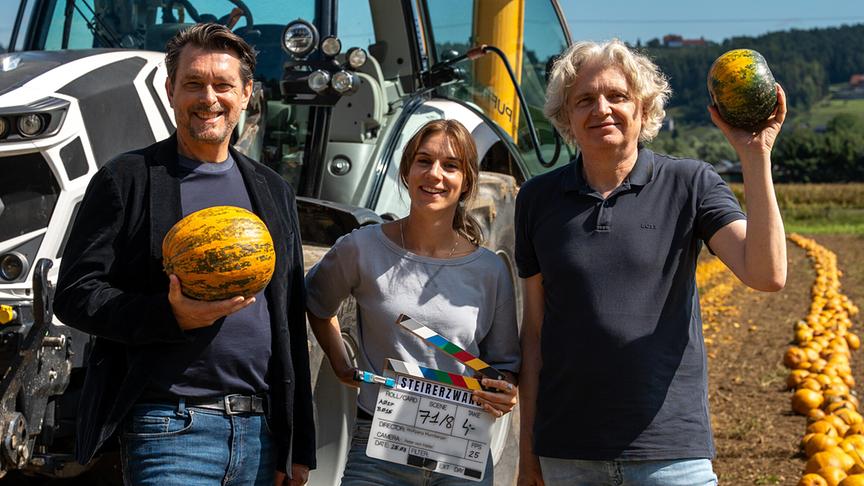 Hary Prinz, Maya Unger und Regisseur Wolfgang Murnberger stehen mit Kürbissen vor einem Traktor auf einem Feld, auf dem eine lange Reihe geernteter Kürbisse liegt.