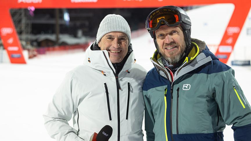Ernst Hausleitner und Hermann Maier stehen in Winterbekleidung im Zieleinlauf vor einer beleuchteten Rennstrecke.