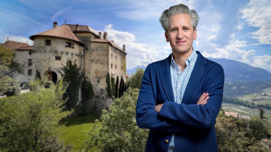 Johann-Philipp Spiegelfeld im blauen Sakko steht vor einem historischen Schloss in einer sonnigen Berglandschaft.