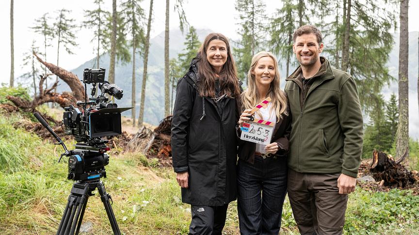 © ORF/Graf Film/Toni Muhr Regisseurin Patricia Frey, Melissa Naschenweng mit einer Filmklappe in der Hand und Ferdinand Seebacher stehen bei einem Filmdreh in einer Berglandschaft neben einer professionellen Kamera.