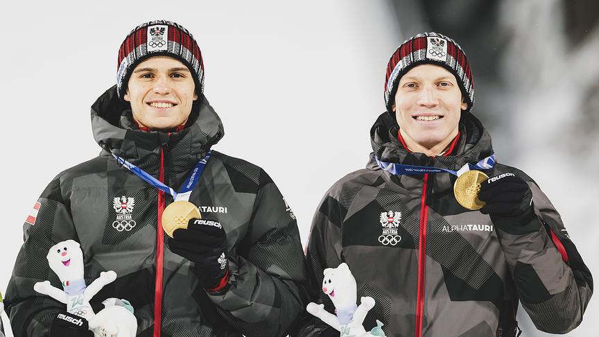Die Goldmedaillen-Gewinner und Olympiasieger Jan Hörl und Stephan Embacher in Teamjacken und Mützen halten Goldmedaillen sowie kleine Maskottchen in die Kamera während der Medaillenzeremonie für das Skispringen Männer Super Team Großschanze im Rahmen der Olympischen Winterspiele in Predazzo.