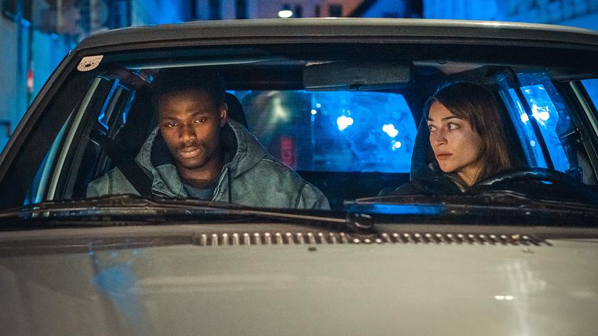 © ORF/ARD Degeto Film/ITV/Anjeza Cikopano Bless Amada und Antonia Moretti sitzen nachts in einem Auto, beleuchtet von blauem Licht einer Polizeieinsatzszene.