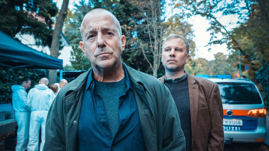 © ORF/ARD Degeto Film/ITV/Anjeza Cikopano Heino Ferch und Robert Stadlober stehen vor einem Polizeifahrzeug, während im Hintergrund Ermittler in weißen Schutzanzügen arbeiten.