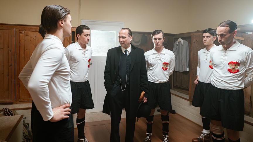 In einer historischen Umkleidekabine stehen mehrere Fußballspieler in alten Trikots um Trainer Hugo Meisl im Anzug, der eine Ansprache hält.