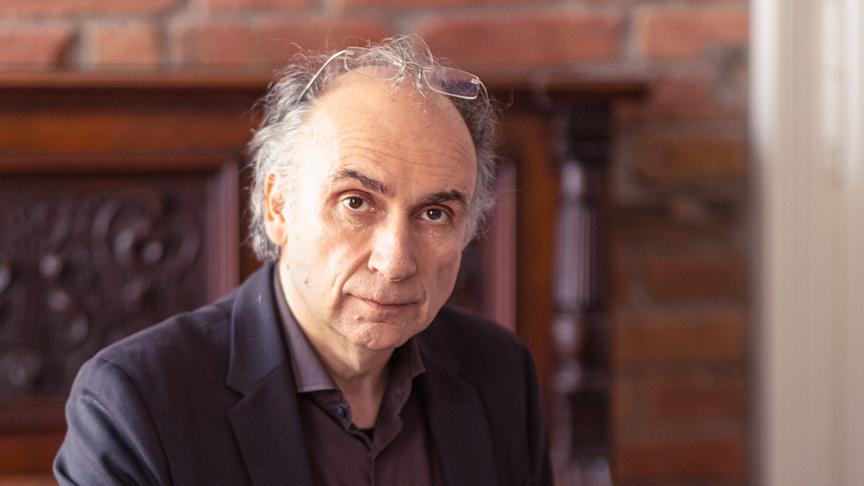 Dimitré Dinev m dunklen Sakko sitzt an einem Holztisch und arbeitet an mehreren ausgebreiteten Dokumenten in einem Raum mit Backsteinwand.