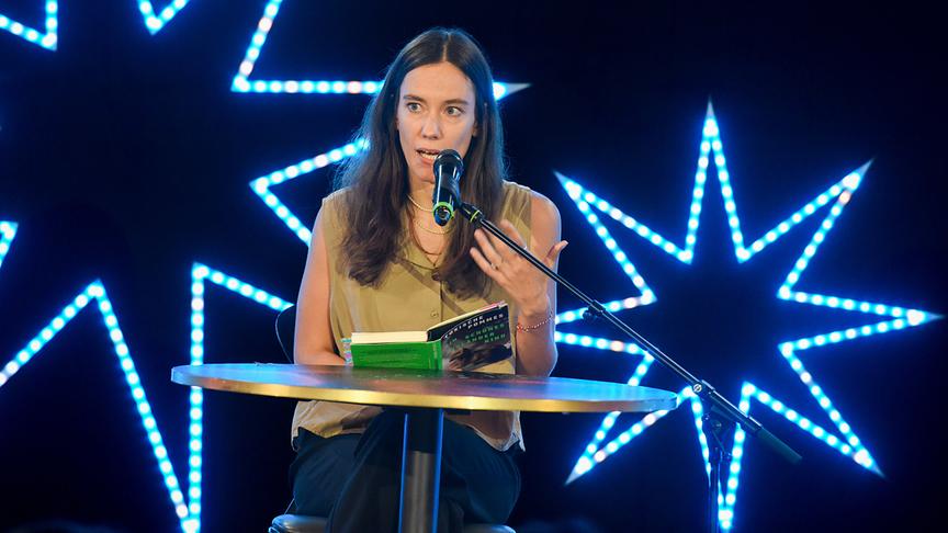 „Toxische Pommes“ sitzt auf einer Bühne an einem kleinen Tisch, liest aus einem Buch und spricht dabei in ein Mikrofon vor einer leuchtenden Sternen-Kulisse.