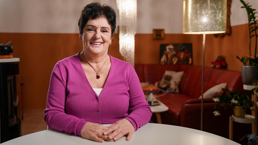 © ORF/Talk TV/Wout Kichler Brigitta, 65, Außendienstmitarbeiterin i.R. aus Niederösterreich, im pinken Cardigan sitzt an einem Tisch in ihrem gemütlich eingerichteten Wohnzimmer.