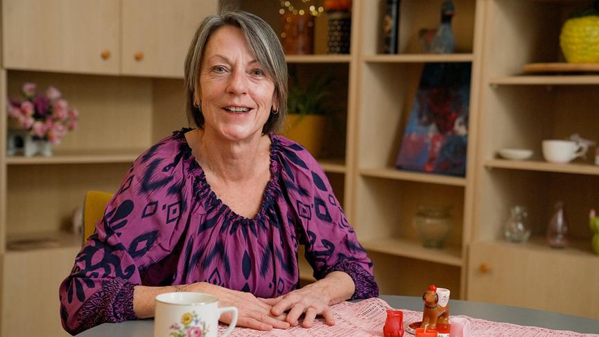 Jutta, 62, Reinigungskraft i.R., Burgenland, sitzt an einem Tisch mit einer Kaffeetasse und kleinen Figuren.