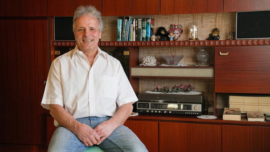 © ORF/Talk TV/Gustl Gschwantner Hans, 66, Tischler i.R. aus der Steiermark, im weißen Hemd und Jeans sitzt lächelnd vor einer holzgetäfelten Wohnzimmerwand mit Büchern, Dekorationen und einer Stereoanlage.