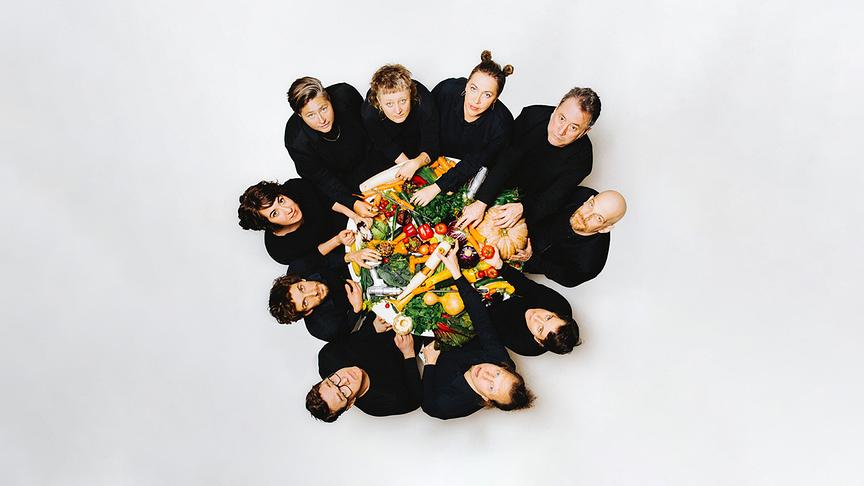 Das Vegetable Orchestra, zehn schwarz gekleidete Menschen, versammeln sich aus der Vogelperspektive um einen Tisch voller buntem Gemüse und blicken nach oben in die Kamera.