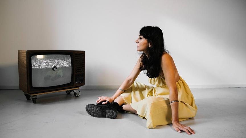 © ORF/Ina Aydogan SAEDI in einem gelben Kleid sitzt in einem schlichten Raum neben einem alten Röhrenfernseher auf dem Boden.