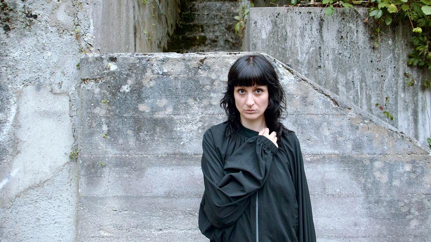 © ORF/Ö1/Michael Poetschko MAIIJA alias Marilies Jagsch mit dunklen, schulterlangen Haaren steht in dunkler Kleidung vor einer verwitterten Betontreppe, umgeben von etwas Grün an den Mauern.