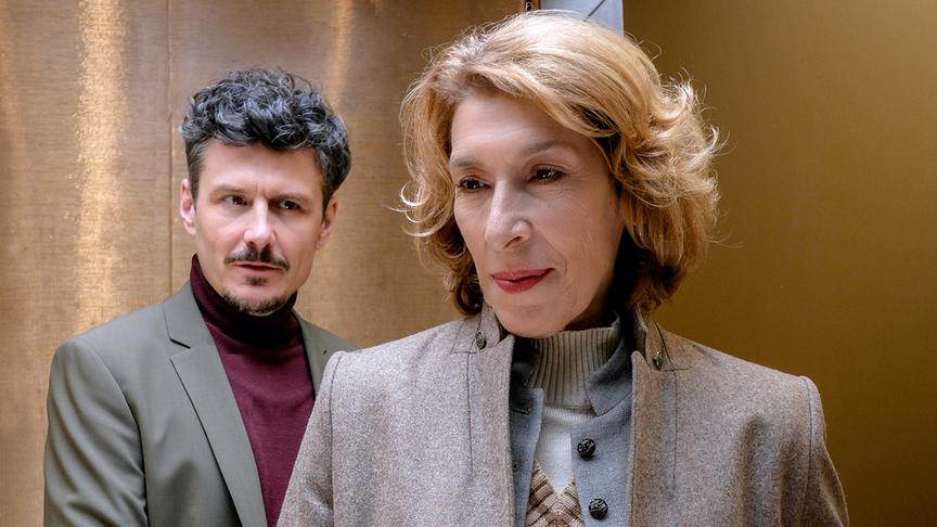 Constanze Laux steht im Vordergrund in Mantel und Strick, während Ricardo Westerhoff im Anzug hinter ihr in einem goldfarbenen Innenraum steht und sie anblickt.