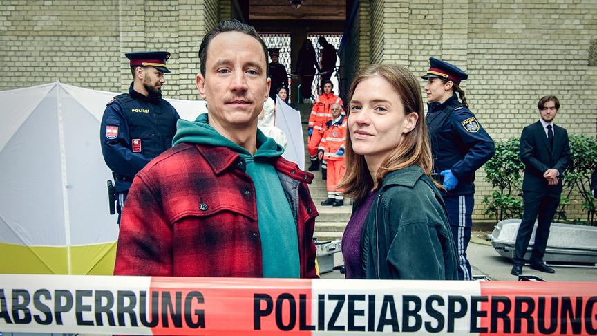 Miriam Fussenegger und Laurence Rupp nehmen Ermittlungen an ihrem ersten Austro-„Tatort“ auf und stehen vor einer abgesperrten Polizeieinsatzstelle, während Polizei‑ und Rettungskräfte im Hintergrund arbeiten.