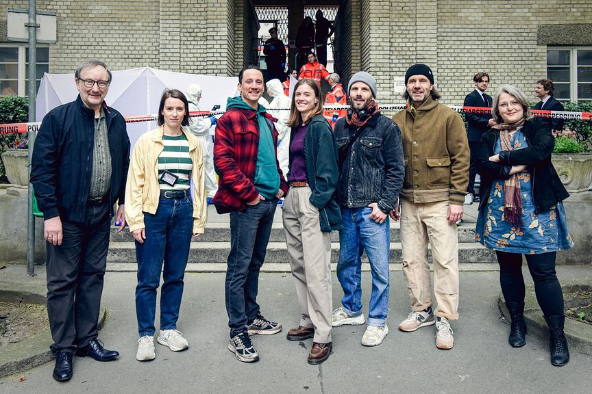 Rainer Bock, Christina Scherrer, Laurence Rupp, Miriam Fussenegger, Regisseur Dominik Hartl, Kameramann André Mayerhofer, Drehbuchautorin Sarah Wassermair stehen am Set zum Austro-„Tatort“  vor einer abgesperrten Einsatzstelle, während Polizei‑ und Rettungskräfte im Hintergrund arbeiten.