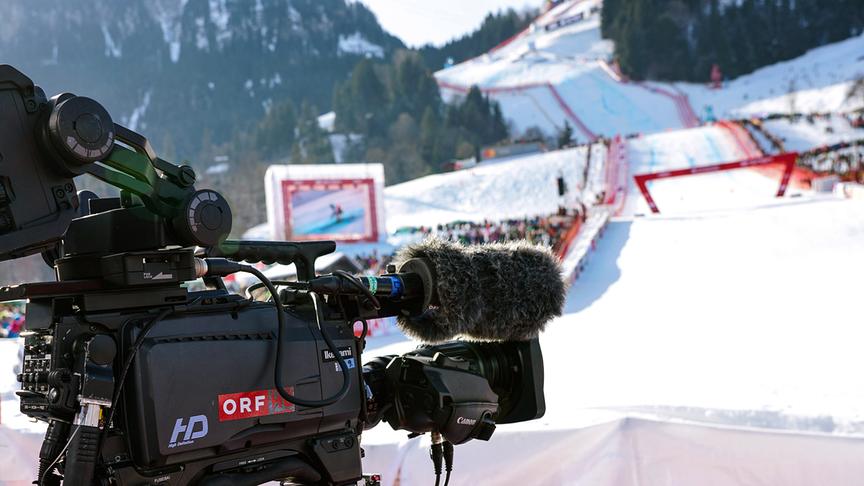 © ORF/Roman Zach-Kiesling Zieleinfahrt in Kitzbühel mit ORF-Kamera mit einer sonnigen Skipiste und Zuschauerinnen und Zuschauern im Hintergrund