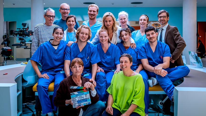 Ein großes Filmteam der ORF-Serie „Pflegeleicht“ posiert in einem Krankenhaus-Set, viele Darstellende tragen blaue medizinische Arbeitskleidung und im Vordergrund ist eine Filmklappe zu sehen. 