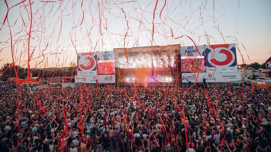 © ORF/Ö3/Mila Zytka Eine große Menschenmenge feiert vor einer Open-Air-Ö3-Bühne am Donauinselfest, während rote Konfettibänder durch die Luft fliegen und die Bühne hell beleuchtet ist.