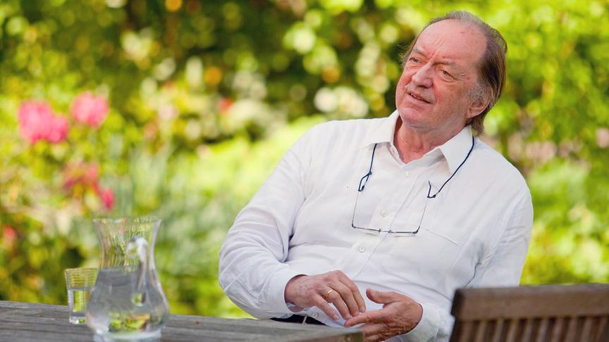 Nikolaus Harnoncourt sitzt beim Interview an einem Holztisch im Grünen, umgeben von Bäumen und Blumen, mit einem Glas und einer Karaffe auf dem Tisch.