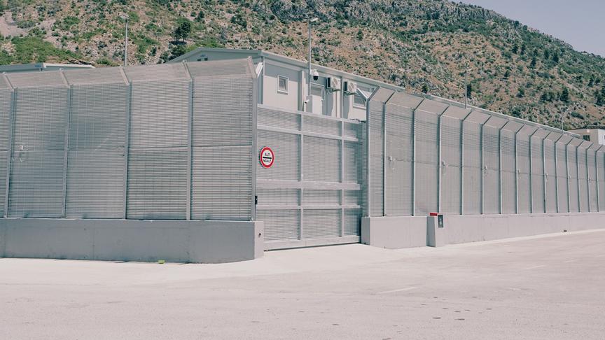 Grenzschutzzaun dahinter ein Gebäude vor einer hügeligen, mediterranen Landschaft